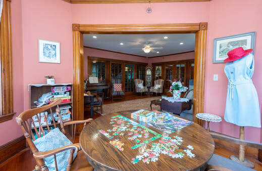 A photo of our dining room. The view is from the table closest to our social room. There are two large oblong tables with a blue tablecloth, white candles and a small pot of yellow flowers in the center of the closest table. Four chairs can be seen and six place settings, each with a floral placemat, red cloth napkin and a setting of silverware, teacup and saucer, and bread plate along with coasters for drinks, tea bags, salt and pepper sakers and dishes of sugar packets. There is one chandelier showing, which has candle-like bulbs on curved arms coming out from a central pendant. There are two windows at the far end. On the right side of the windows sits a fireplace with a mirror set into the wood framing of the mantel and surround. On the left side of the window is a wooden china hutch. Lace valances hang from the top of the large, bright windows.