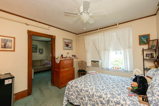 Photo of a bedroom suite. Pictured are a bed and dresser near a double window. There is a doorway next to the dresser and a couch in the next room. Personal items of an elderly woman are staged around the room.