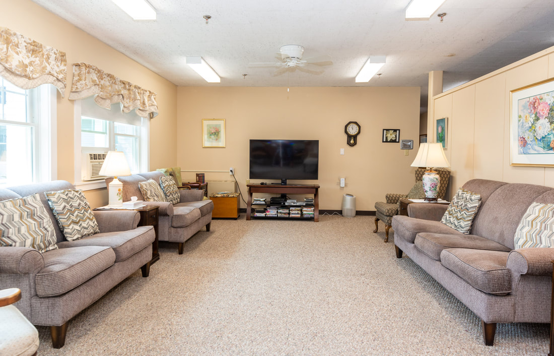 A photo of our front parlor. It is painted a muted pink and there are three visible windows overlooking Lothrop St. There is a soft pink high-backed chair to the right.  On the right side of that chair is a piano. On the right side of the piano are two high-backed chairs with a blue and green striped pattern. Above and behind those chairs is a large golden mirror hanging on the wall. In a small alcove with two visible windos sits a round table and two chairs along with two large potted plants. On the far left side of the photo is a small couch and coffee table adorned with a long lace runner. At the center of the room is a large muted pink and blue floral carpet.